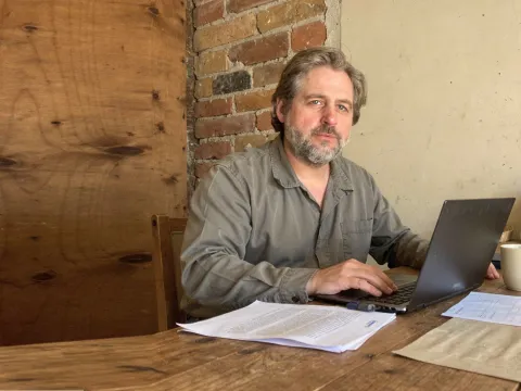 Sebastian Klein an einem Holztisch vor einer Backsteinwand, vor sich ein Laptop und Unterlagen